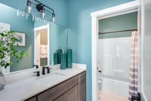 Bright, modern bathroom with teal walls, a large mirror, white sink, brown cabinets, and a view into an adjoining room with a shower and striped curtain.