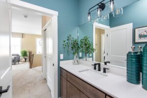 Modern bathroom with a double vanity, large mirror, teal walls, and a view into an adjoining beige bedroom.