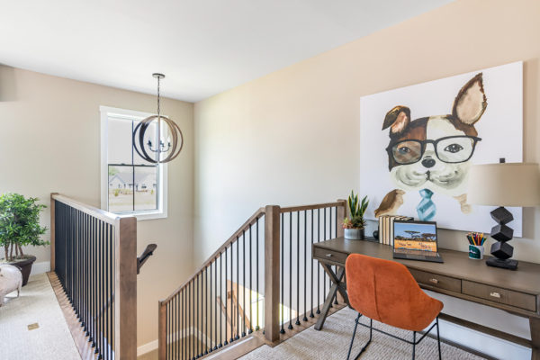 A well-lit home office space with a wooden desk, an orange chair, and framed artwork of a dog wearing glasses and a tie. a round light fixture hangs near the window.