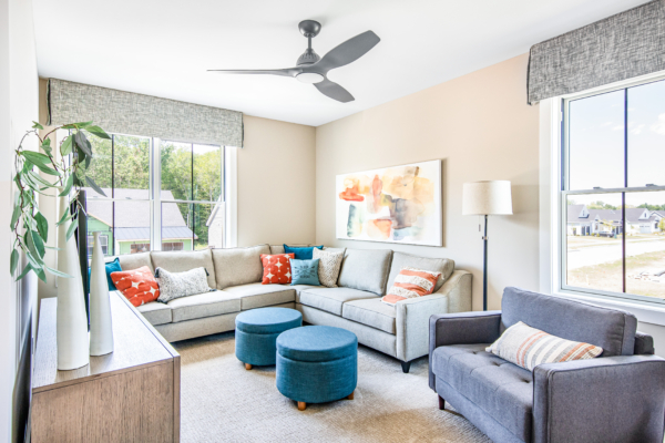 Bright, contemporary living room with a large sectional sofa, two blue ottomans, and a wall-mounted artwork above a couch under a ceiling fan.