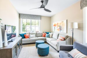 Bright living room with a gray sectional sofa, blue ottomans, beige walls, large windows with blinds, and a ceiling fan.