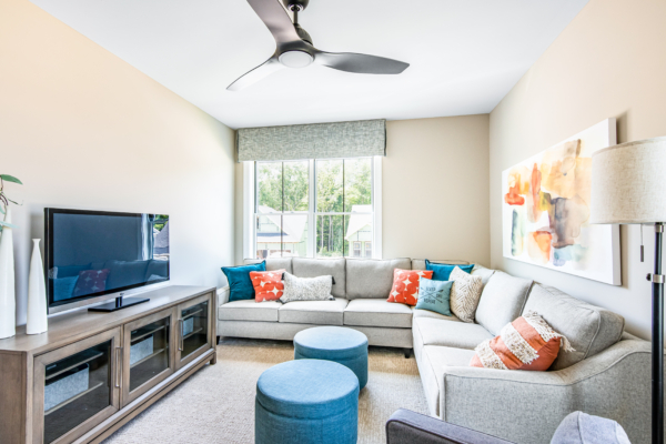 Bright living room with a large sectional sofa, colorful cushions, a ceiling fan, and a flat-screen tv on a wooden stand. two round blue ottomans and large windows are also visible.