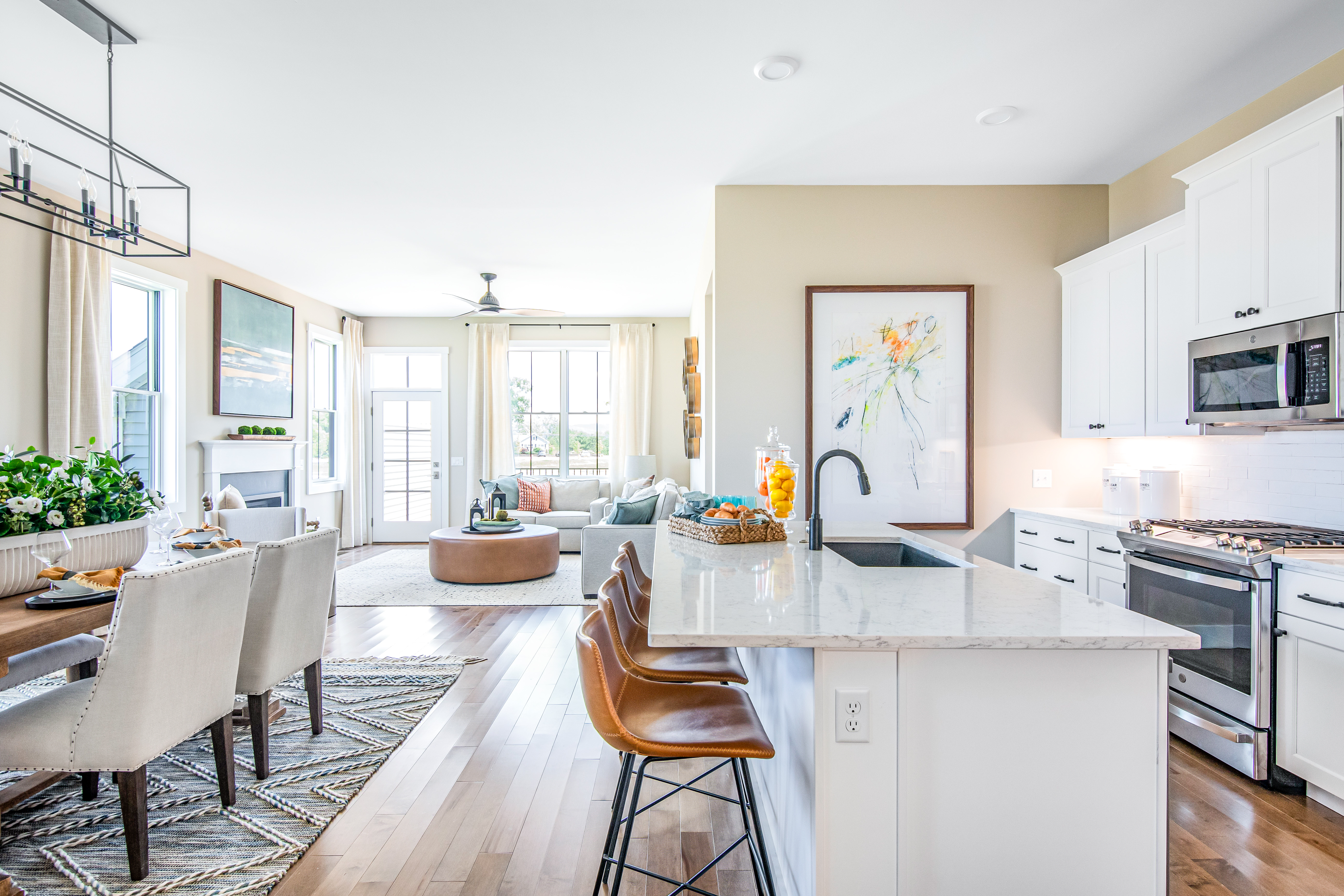 Bright, open-plan kitchen and living area with modern white cabinets, stainless steel appliances, a central island with bar stools, a dining area, and a cozy seating area leading to a sunny balcony.