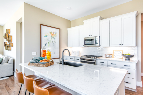 Modern kitchen interior with white cabinetry, marble countertops, and stainless steel appliances, decorated with a fruit basket and wall art.