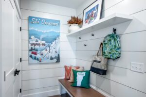 A modern entryway featuring white shelves, a bench with decorative pillows, hooks with a backpack and bag, and a colorful "ski vermont" poster on the wall.