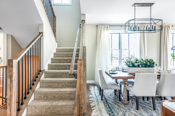 Modern dining room with a staircase leading upstairs, featuring a wooden table, white chairs, and a hanging chandelier.