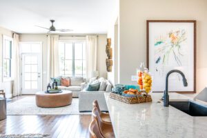 Bright, modern kitchen with a marble countertop and living room view, featuring neutral decor, a large fruit-filled centerpiece, and an abstract wall art.