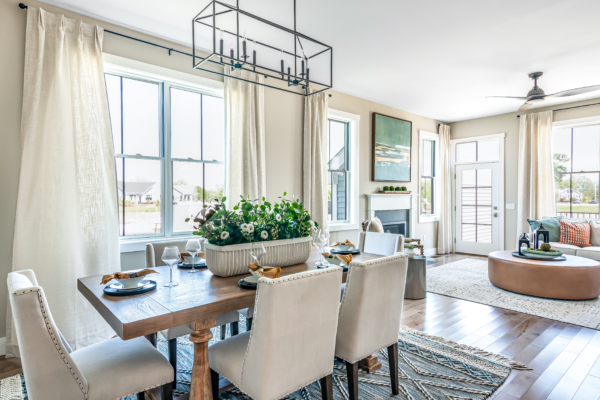 Modern dining room featuring a wooden table with chairs, large windows with sheer curtains, and an adjacent living area with a circular rug and cream sofa.