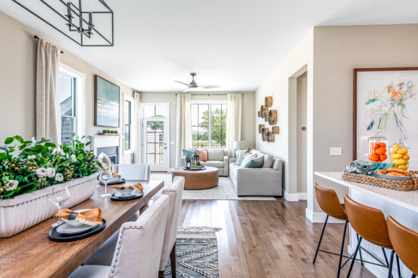 A bright, modern dining and living room with a wooden table set for a meal, a gray sofa, large windows, and colorful wall art.