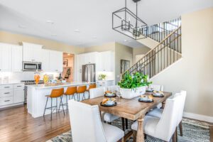 Bright, modern kitchen with white cabinets and a central island with orange stools, connected to a dining area with a wooden table set for a meal.