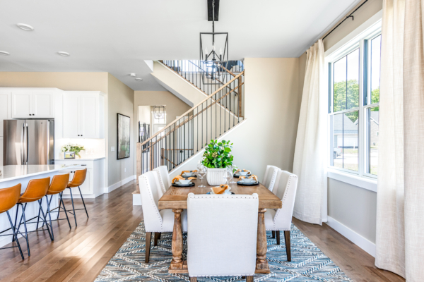 Bright, modern dining area with a white table set for four, adjacent to a kitchen with bar stools, under a staircase and ample natural light from large windows.