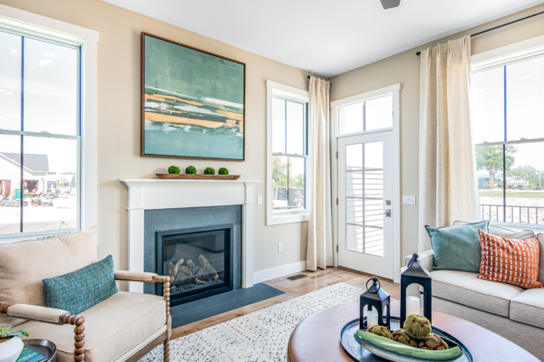 A bright, modern living room with a fireplace, large windows, a beige sofa, colorful cushions, and a painting above the mantel.