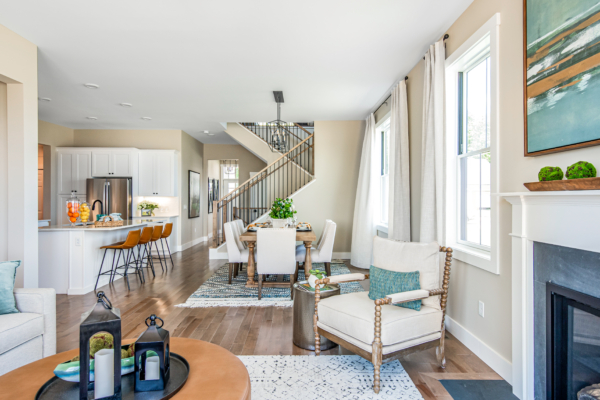 Bright, open-plan living area with a kitchen, dining space, and seating by a fireplace, featuring large windows and modern decor.