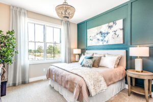 Modern bedroom featuring a teal accent wall with abstract art, plush beige bedding, a crystal chandelier, and a large window with curtains.