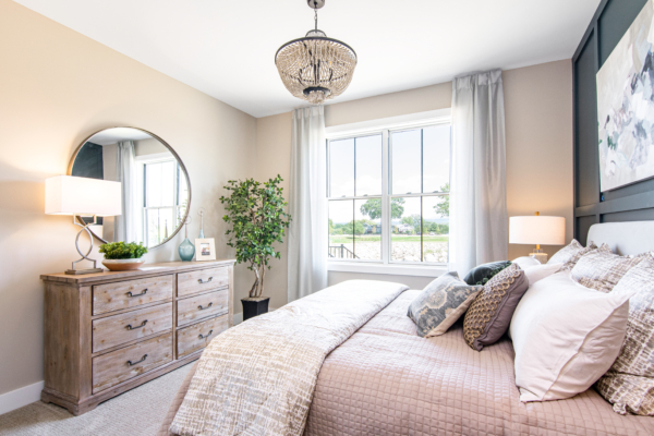 Bright and airy bedroom featuring a large bed with pastel bedding, a round mirror, a dresser, and large windows with gray curtains.