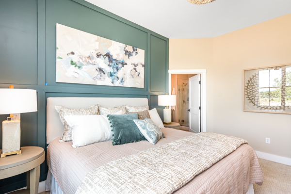 A well-lit bedroom featuring a queen-sized bed with beige and green pillows, a large abstract painting above it, side tables with lamps, and an open door.
