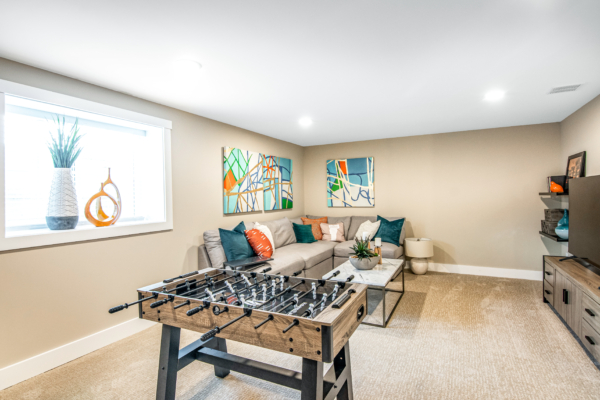 Modern basement living area with a foosball table, l-shaped sofa, colorful cushions, abstract wall art, and a window displaying outdoor greenery.