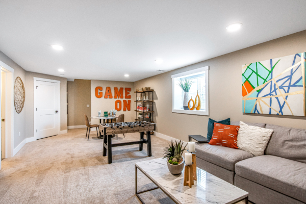 A modern basement living space featuring a "game on" wall decor, sectional sofa, a glass coffee table, bright artwork, and ambient lighting.