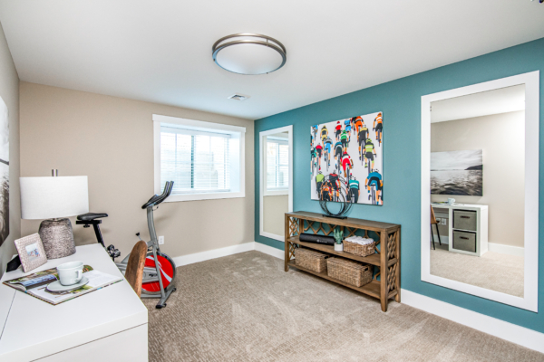 A modern room with exercise equipment, a desk with a lamp, and colorful wall art, featuring neutral carpets and light blue walls.