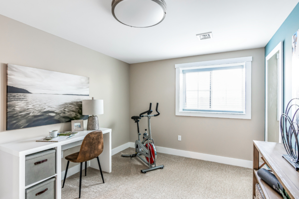 A modern home office with a desk, chair, exercise bike, and a large window with a picturesque view, complemented by neutral-toned walls and decor.