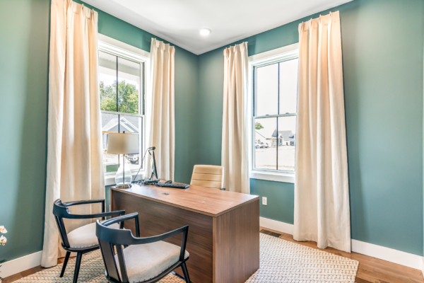 A well-lit home office with teal walls, tan curtains, a wooden desk with a laptop, a beige chair, and a blue armchair near a window.