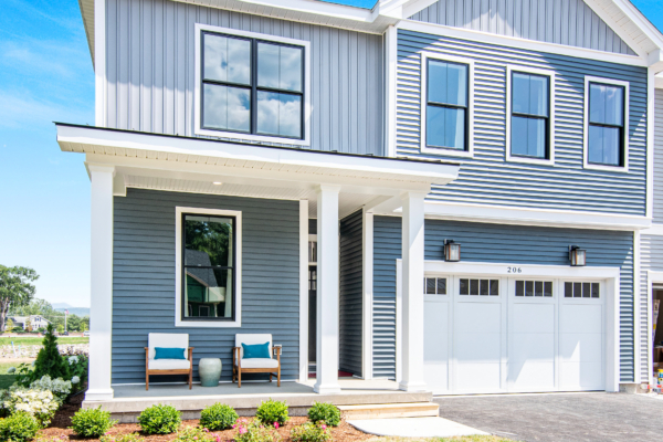 Modern two-story house with blue siding, white trim, and large windows, featuring a front porch with two chairs and landscaped front yard.