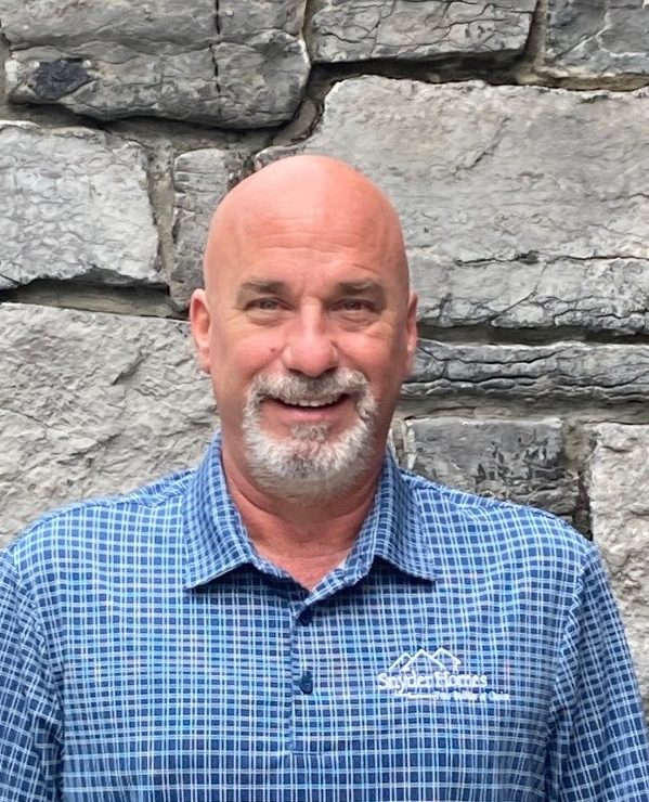 A smiling bald man wearing a blue checkered shirt standing in front of a stone wall.