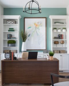 A stylish home office with a wooden desk, white built-in shelves, and a large abstract painting above the desk, against a light teal wall.