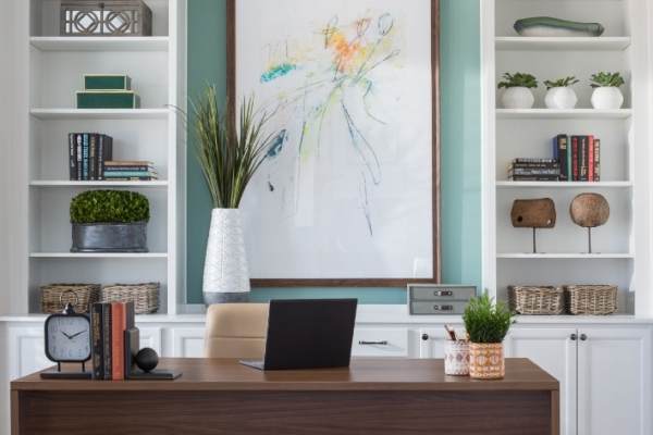 A stylish home office with a wooden desk, white built-in shelves, and a large abstract painting above the desk, against a light teal wall.