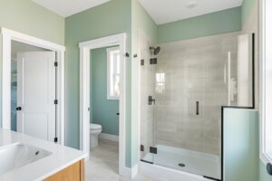 Modern bathroom featuring a glass shower enclosure, white bathtub, and pale green walls, with a view into an adjoining room.