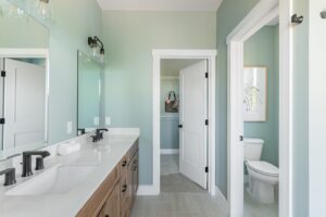 A modern bathroom featuring dual sinks, large mirrors, white countertops, wooden cabinets, and a separate toilet area with an open door. light green walls and bright lighting enhance the clean aesthetic.