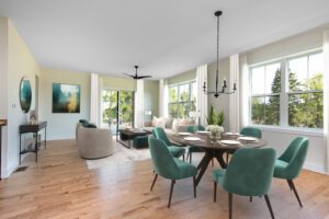Spacious living and dining room with large windows, teal dining chairs, a wooden table, and a separate sitting area with a sofa.