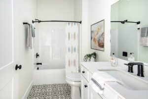 Modern bathroom with a white bathtub, patterned floor tiles, white walls, and a vanity mirror. towels hang on black racks, and there's a plant on the counter.