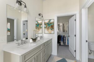 Modern bathroom with double sinks, mirrors, and artwork, connected to a walk-in closet containing clothes and accessories.