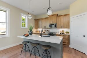 Modern kitchen with light wood cabinets, stainless steel appliances, white countertops, a central island with stools, pendant lights, and light walls.