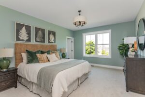 A serene bedroom featuring a queen-sized bed with white and green bedding, botanical art above the headboard, and a soft green wall color.