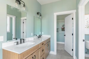 A modern bathroom with dual sinks, wooden cabinets, and a sea green wall, featuring bright lighting and an open closet door.