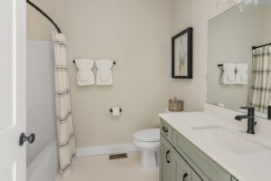 Modern bathroom with beige walls, featuring a green vanity, white sink, toilet, and a shower with a striped curtain. towels are neatly hung and a framed picture decorates the wall.