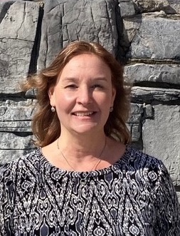 A middle-aged woman smiling, with shoulder-length blond hair, dressed in a patterned blouse, standing in front of a rugged stone wall.