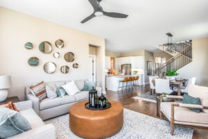 A bright, modern living room with white sofas, a round leather ottoman, and a dining area in the background, decorated with multiple round mirrors on the wall.