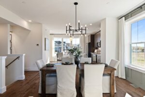 Bright dining area with a wooden table, six chairs, modern chandelier, and kitchen in the background.