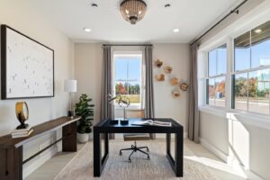 Modern home office with a black desk, art on the wall, large windows, and natural light streaming in.