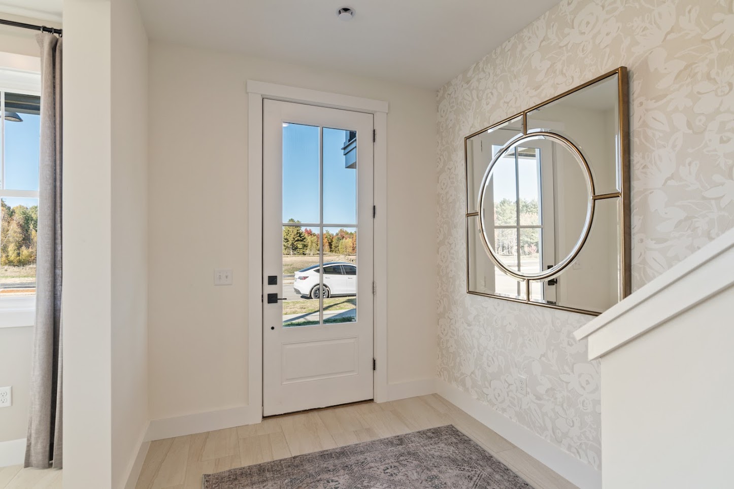 Bright entryway with a glass door, decorative mirror, floral wallpaper, and a gray rug.