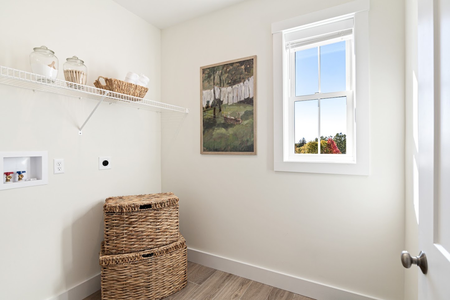 Laundry room with two woven baskets, wall art, a shelf with jars, and a window with sunlight streaming in.