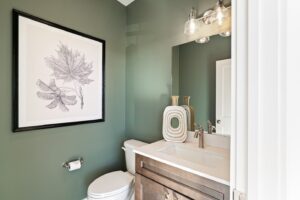 A small bathroom with green walls, a toilet, sink, large leaf artwork, and modern light fixtures.