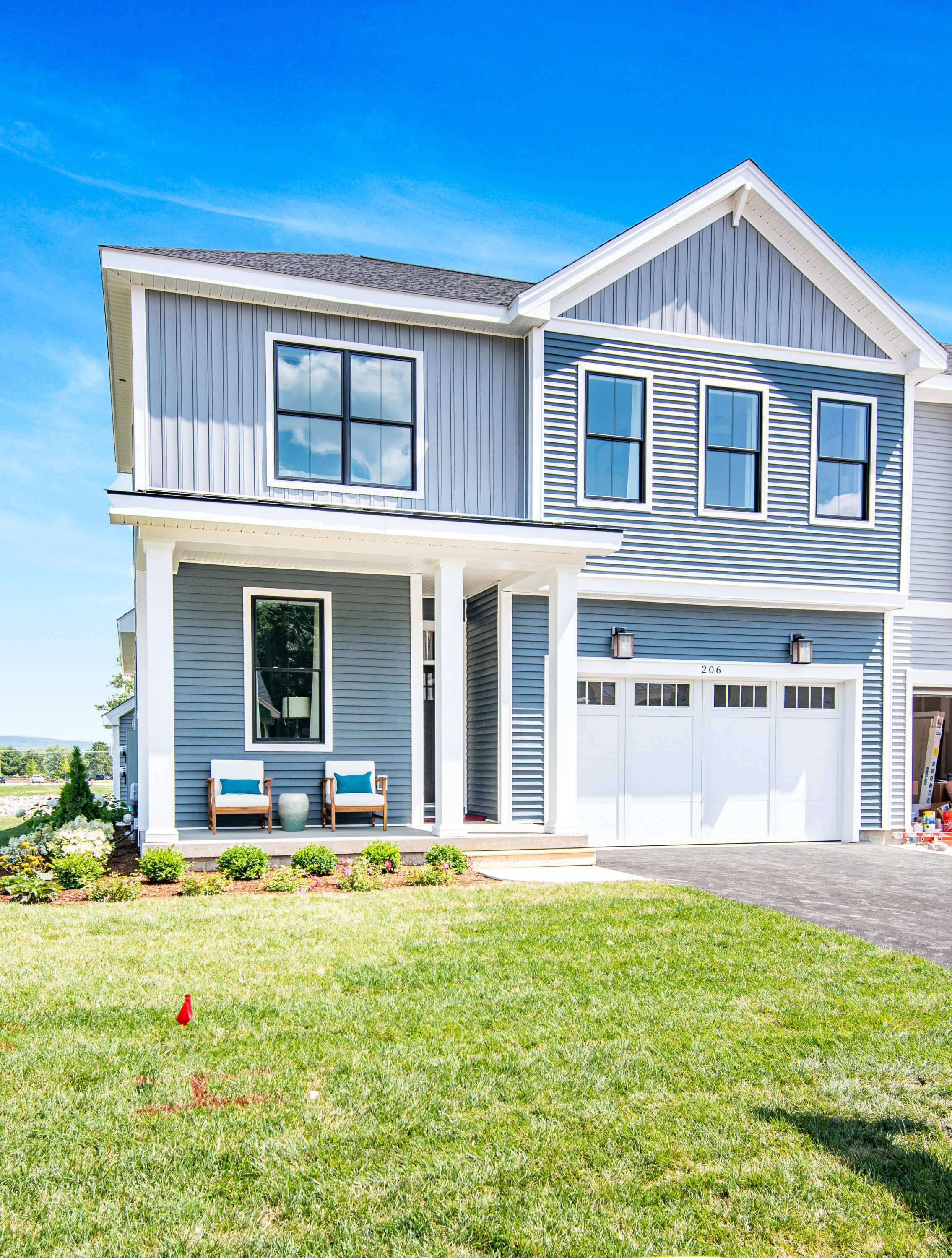 Two-story blue house with white trim, featuring a front porch with two benches and a potted plant. There is a driveway leading to a two-car garage on the right. Bright green lawn in front.