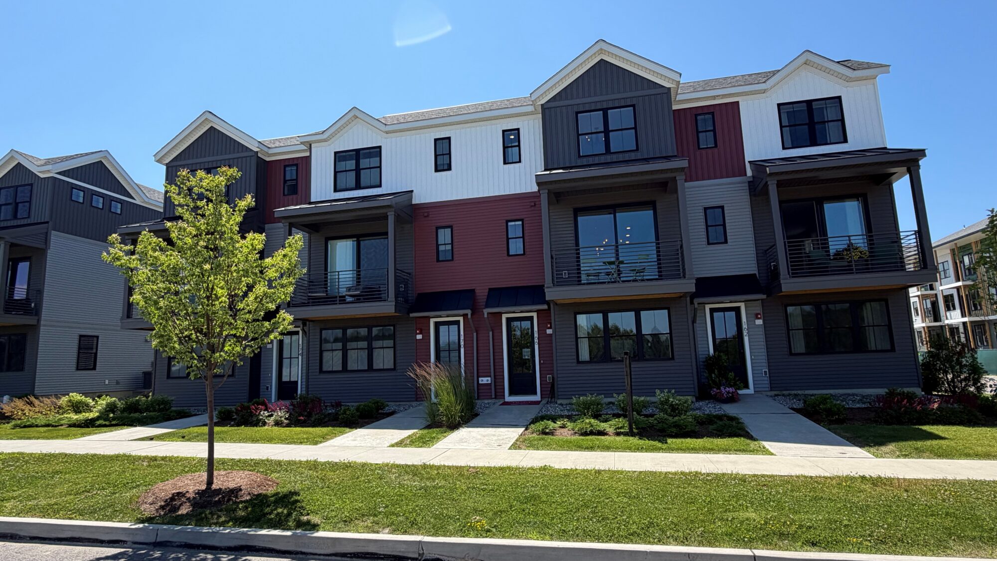 Modern three-story townhouse complex with balconies, a green lawn, and a young tree in front under a clear sky.