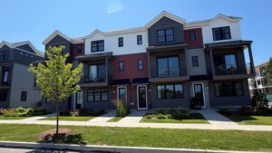 Modern three-story townhouse complex with balconies, a green lawn, and a young tree in front under a clear sky.