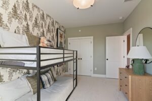 Kids’ bedroom with a metal bunk bed, dresser with lamp, and tree-patterned wallpaper on one wall.