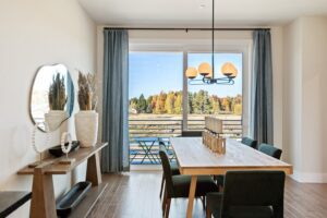 Modern dining room with wooden table, green chairs, large window, and autumn trees visible outside.